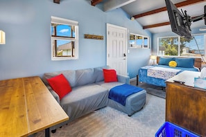 Una habitación acogedora con una mesa de madera, un sofá gris con cojines rojos, una cama azul y un televisor montado.