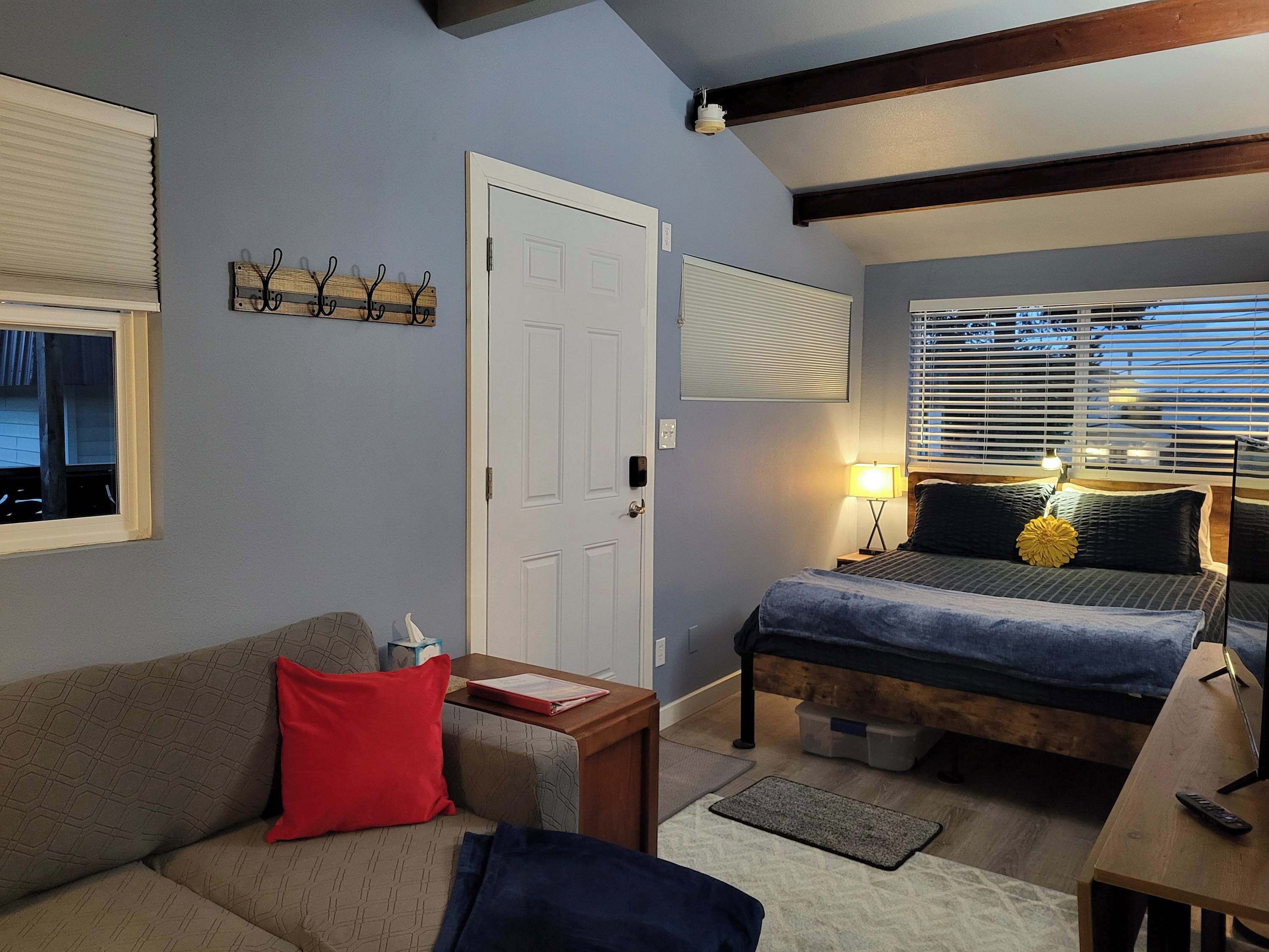 A cozy room with a bed, a sofa, a small table, and a desk. There is a window with blinds, a door, and a wall-mounted light fixture.