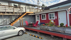 A two-story building with a staircase leading to the upper floor. There is a red building adjacent to it with a white door and windows. A car is parked in front of the building, and there are red chairs and a table outside. - Studio with covered deck and Birch Bay View (Blaine)