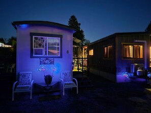 Ein kleines, beleuchtetes Haus mit Veranda, zwei Stühlen und einem Blumentopf.