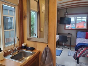 Ein Zimmer mit Holztür, Waschbecken mit Spiegel, Bett mit Patchworkdecke, Schreibtisch mit Stuhl und Fernseher an der Wand.