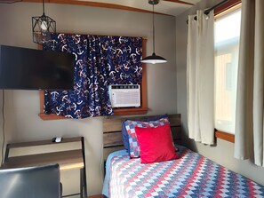 Ein Schlafzimmer mit Bett, Schreibtisch, Stuhl, einem Fenster mit Vorhängen und einer Pendelleuchte.