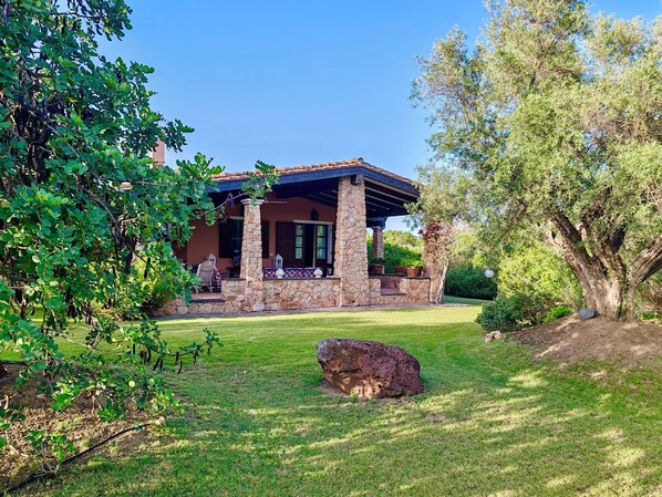 A stone house with a covered patio, surrounded by lush greenery and a large rock in the foreground.