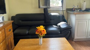 En stue med en sort lædersofa, et træsofabord med en vase blomster, et træskab, et fjernsyn og et køkkenområde med en vask og skabe.