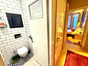 Ein kleines Hotelzimmer mit weiß gefliester Wand, einem Spiegel in Schwarzrahmen, einer weißen Toilette und einem gemusterten Boden.