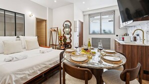 A modern hotel room with a large bed, a dining table set for two, a kitchen area, and a TV mounted on the wall.