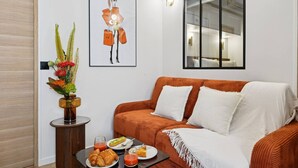 A modern living room with a brown sofa, a small table with breakfast items, a framed artwork, and a vase with flowers.
