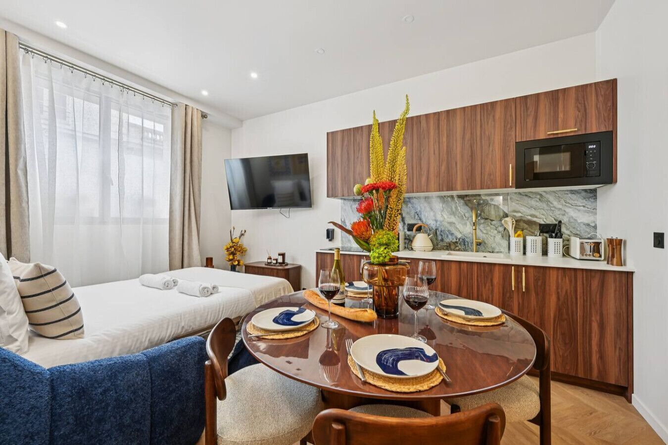 A modern hotel room with a bed, a sofa, a dining table set with plates, glasses, and a vase of flowers.