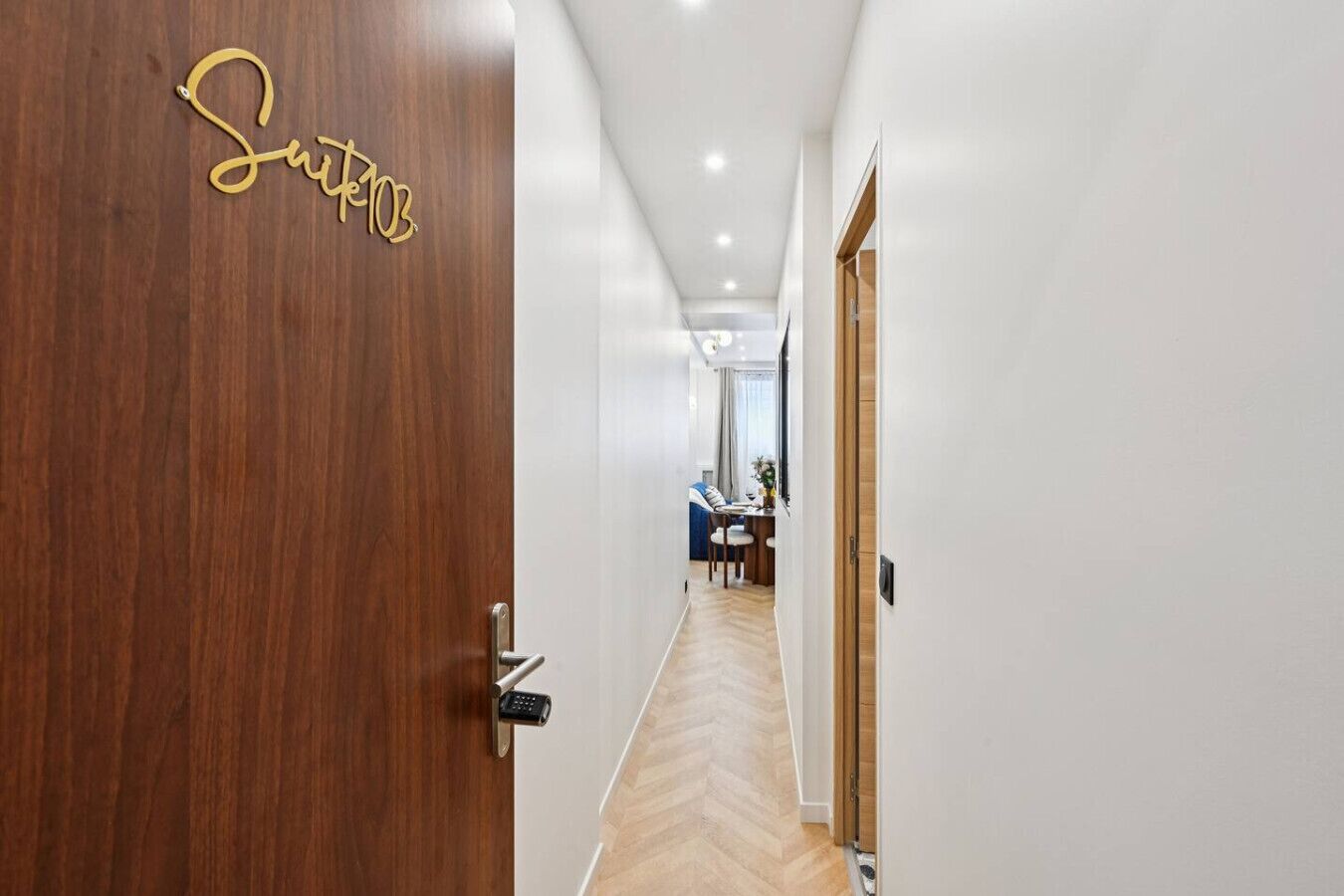 Un couloir mène à une porte en bois portant l’inscription « Suite 103 », qui donne sur une pièce contenant un bureau et une chaise.