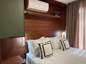 Een hotelkamer met een houten hoofdeinde, een bed met witte beddengoed en zwart afgewerkte kussens, een ingebouwde airconditioner en een kleine potplant op het hoofdeinde.
