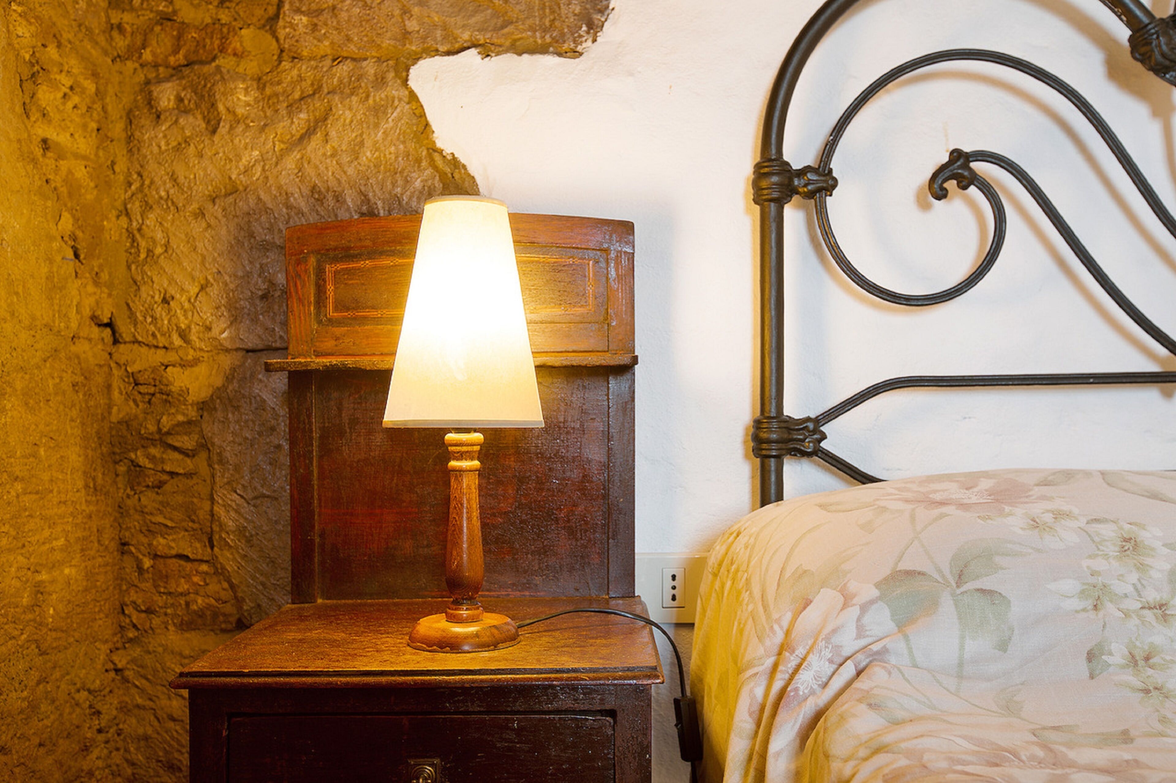 A wooden bedside table with a lamp, a metal headboard, and a floral bedspread.