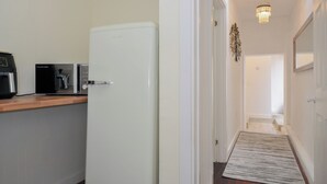 A compact kitchen with a refrigerator, coffee maker, and toaster. A narrow hallway leads to a door with a light fixture above. - Stargazy (Tintagel)