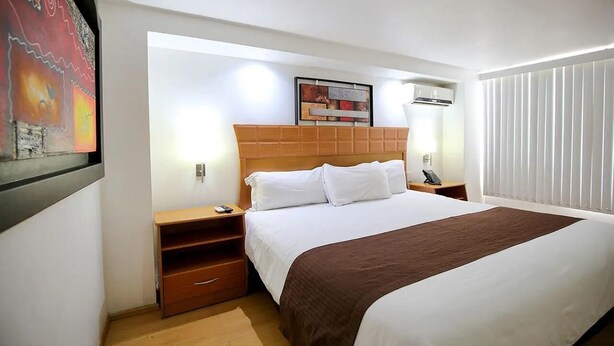 Een hotelkamer met een groot bed, een houten hoofdbord, nachtkastjes, een venster met jaloezieën en een ingelijste foto aan de muur.