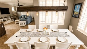 A dining area with a large table set for four, surrounded by white chairs, with a rustic wooden beam above. - Pet-Friendly 4 bed Home w/ Pool Access, Patio & Garage – Walk to Dining! (Gilbert)