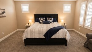 Un dormitorio con cama, dos mesitas de noche, una silla y un ventanal con persianas.