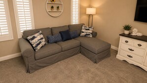 A living room with a sectional sofa, a lamp, a TV, and a white dresser. - Pet-Friendly 4 bed Home w/ Pool Access, Patio & Garage – Walk to Dining! (Gilbert)