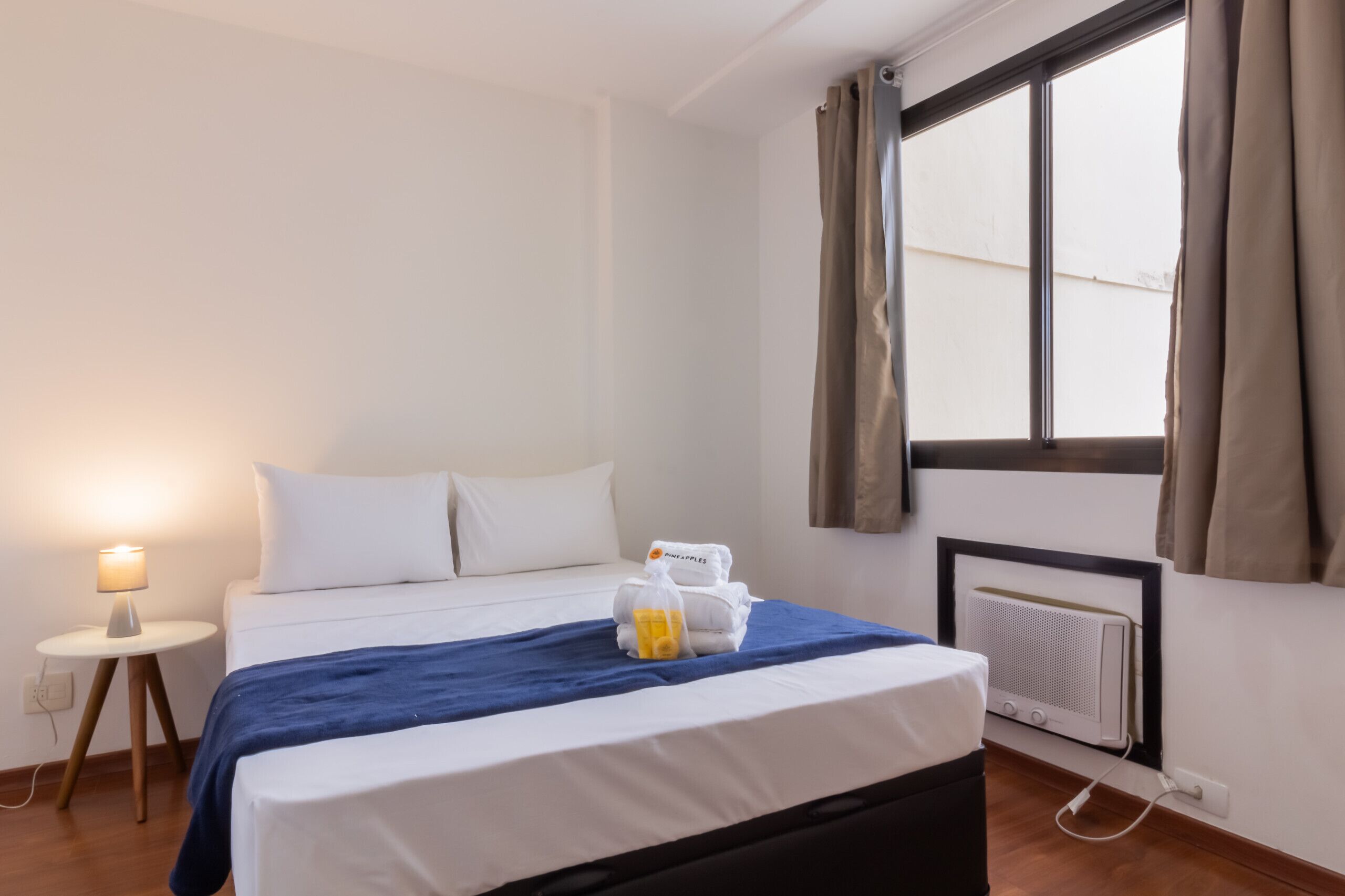 Ein ordentlich bezogenes Bett mit blauer Decke, weißen Kissen und einem kleinen Tisch mit Lampe daneben.