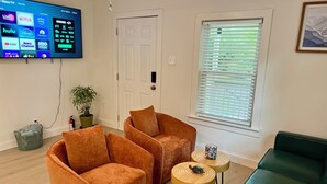 Una sala de estar con dos sillones naranjas, un sofá verde, una mesita de madera con una figurita de tortuga y un televisor mostrando servicios de streaming.