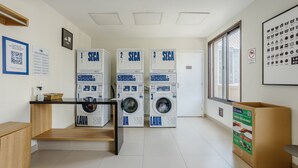 Ein Waschraum mit vier Waschmaschinen, die mit 'SECA' und 'LAVA' beschriftet sind. An den Wänden befinden sich Hinweisschilder, ein Fenster mit Ausblick und ein Karton auf dem Boden.
