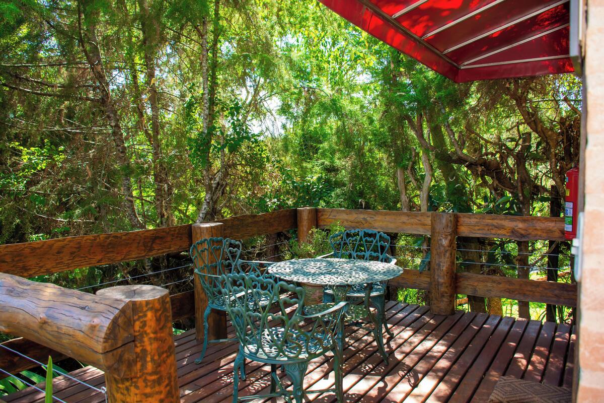 Une terrasse en bois avec une table et des chaises en métal, entourée d’arbres.