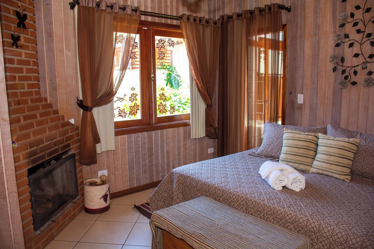 Une chambre avec une cheminée en briques, un lit recouvert d’une couvre-lit à motifs et des rideaux aux fenêtres.