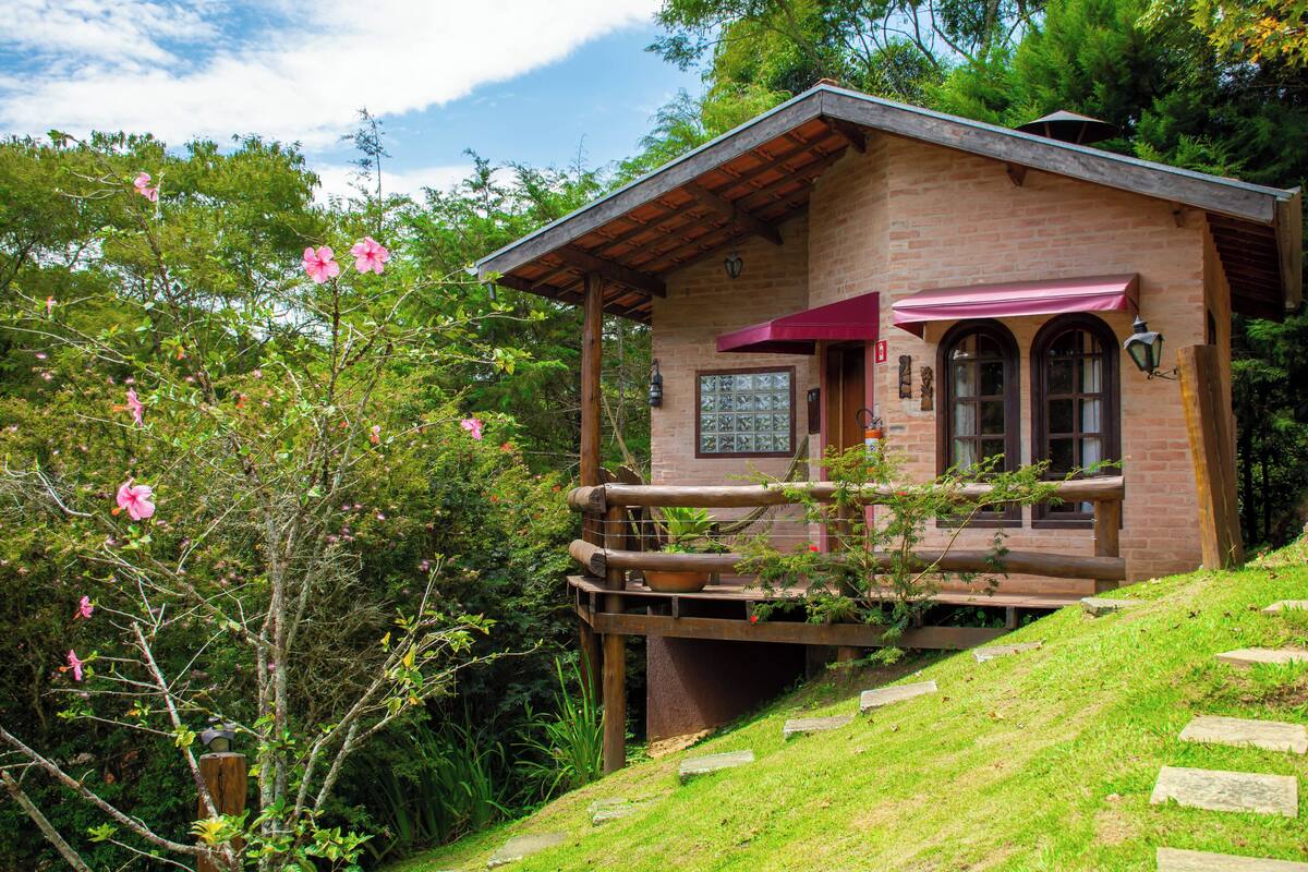 Une maison de campagne en briques, dotée d’un balcon en bois, entourée d’une végétation luxuriante et de plantes en fleurs.