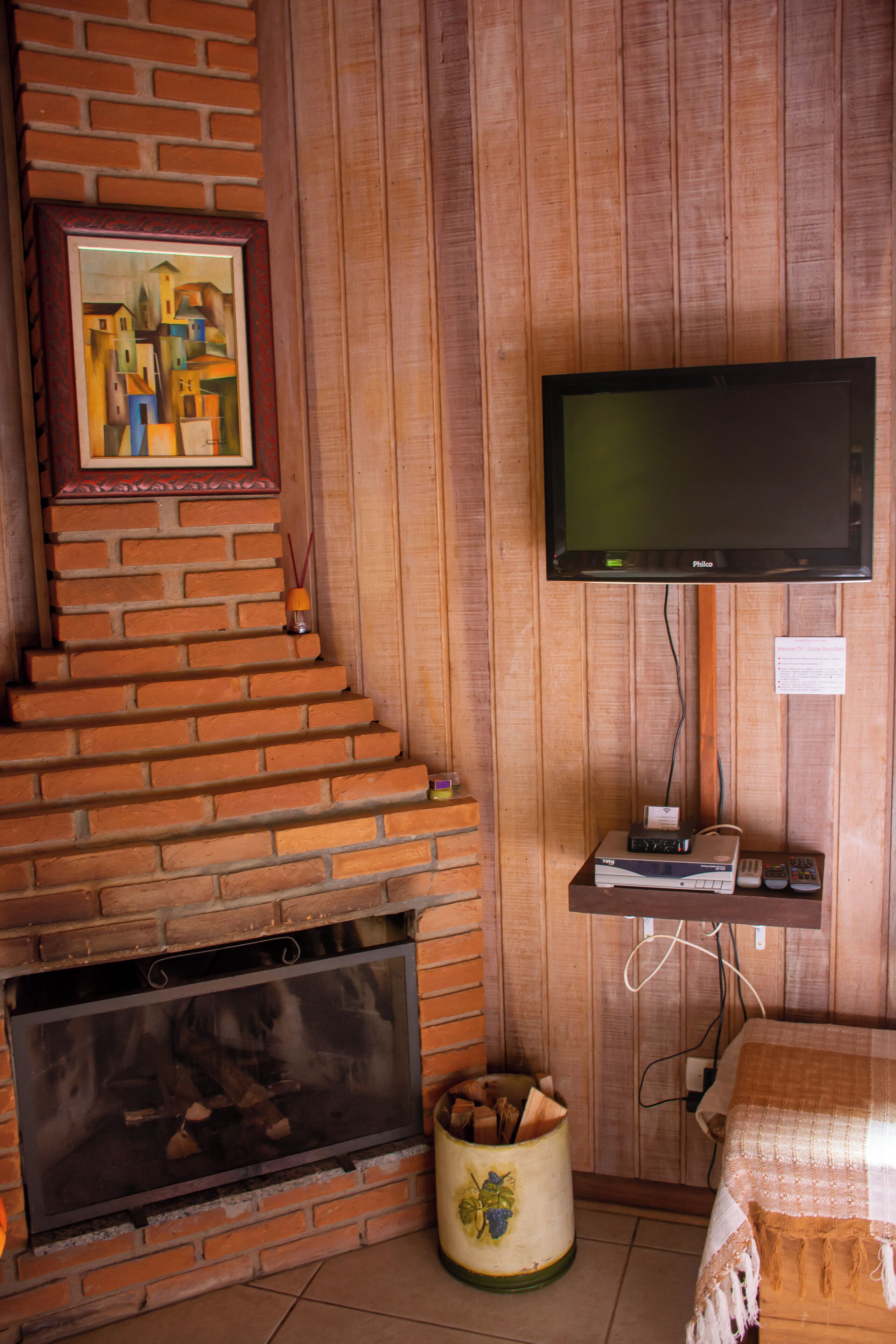 Quarto com lareira de tijolos, televisão de tela plana presa a uma parede de madeira, uma pequena prateleira com dispositivos eletrônicos e uma cama aconchegante com um cobertor.