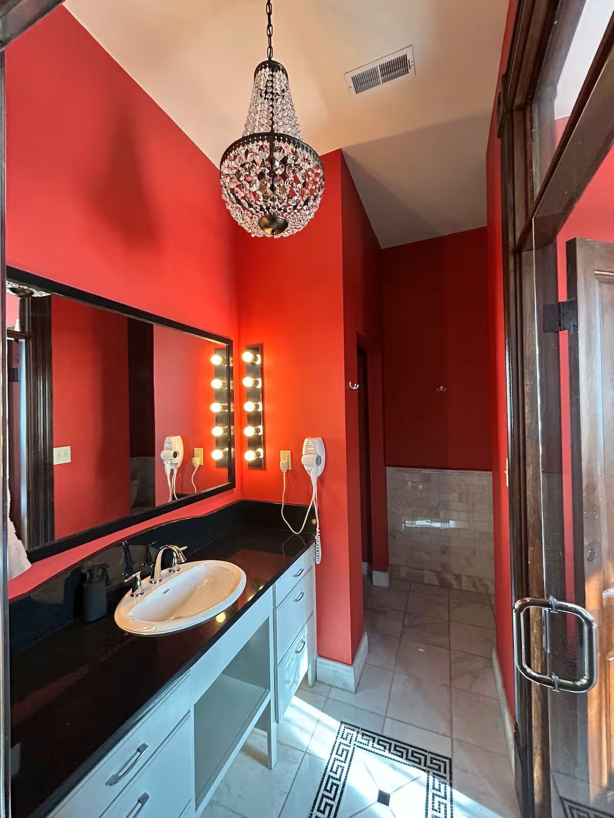 Un bagno moderno con un piano d'appoggio nero, uno specchio grande illuminato, un lampadario e una parete rossa.