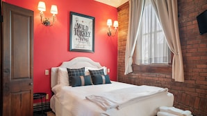 A hotel room with a red wall, a white bed with multiple pillows, a framed picture on the wall, and a window with curtains. - Southern Charm and Comfort on the Bourbon Trail- Trail Suites Inn, Suite C (Lawrenceburg)