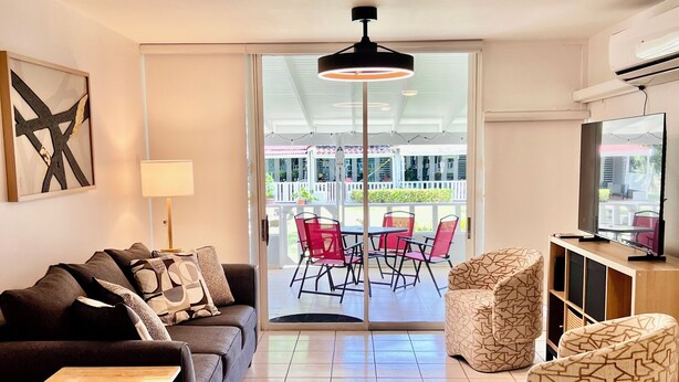 Ein modernes Wohnzimmer mit einer Couch, einem kleinen Tisch, einem Sessel und einer TV-Konsole. Ein Balkon mit roten Stühlen und einem Tisch ist ebenfalls vorhanden.