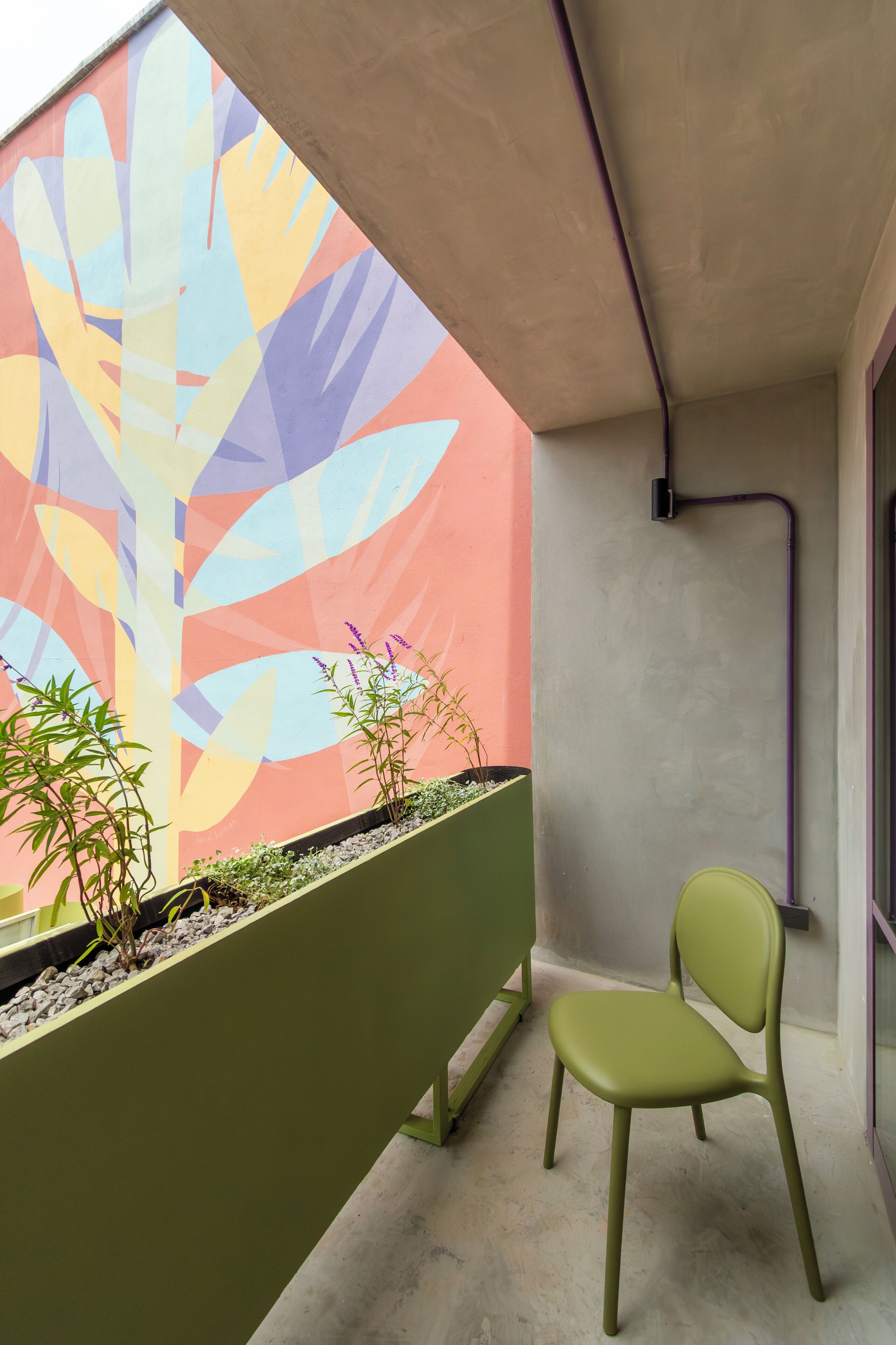 Un balcón con un mural de colores, una jardinera verde con plantas y una silla verde.