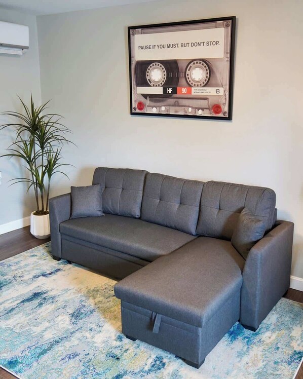 A grey sectional sofa with a matching ottoman, a blue and white patterned rug, and a framed poster on the wall. - Hidden Sanctuary (Hagerstown, MD) (Hagerstown)