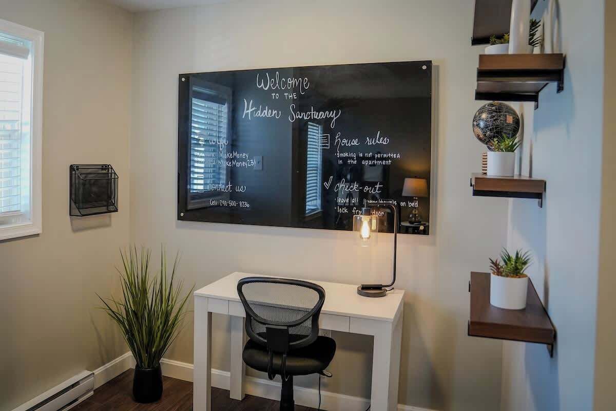 A room with a desk, a potted plant, and a chalkboard displaying house rules.