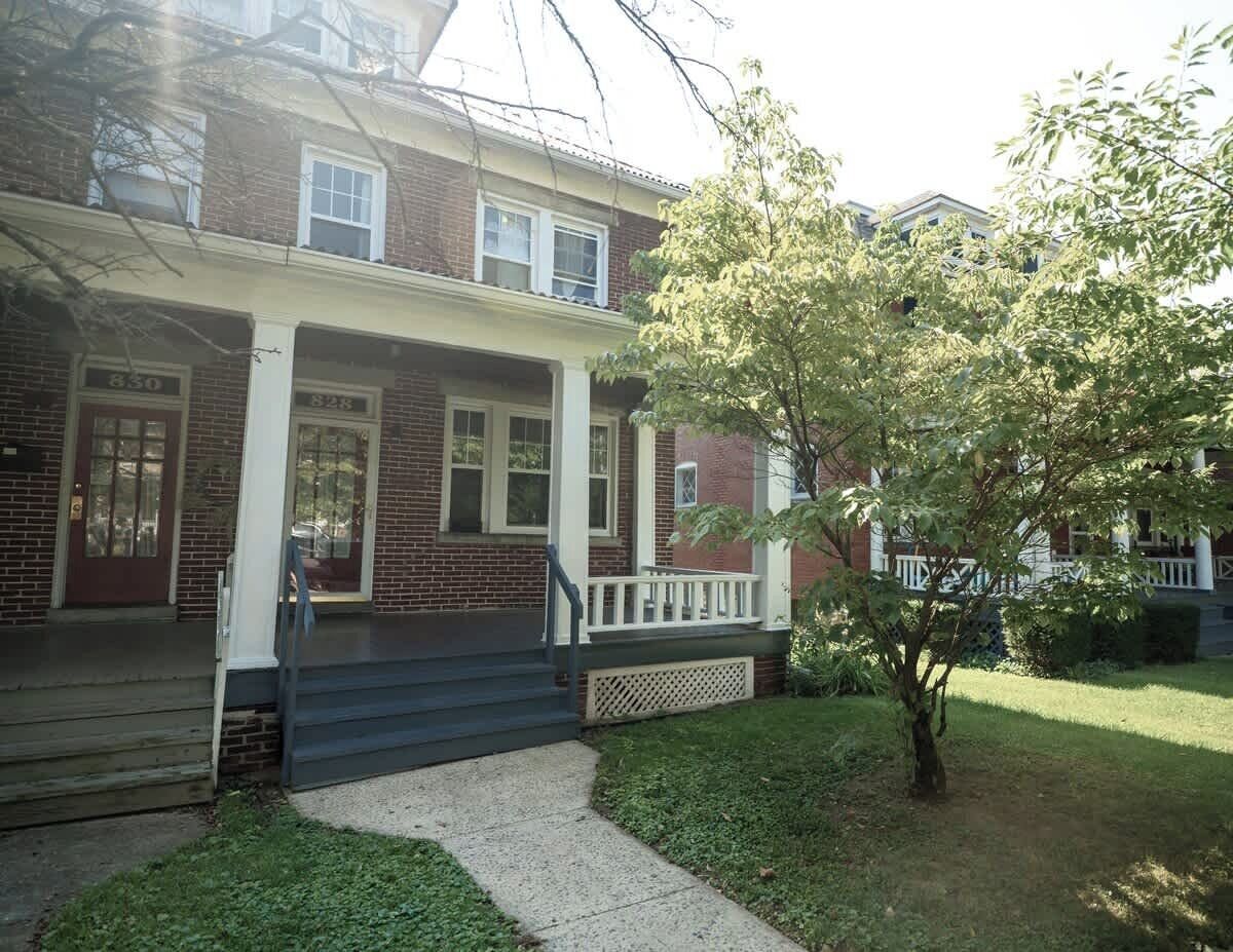 A two-story brick house with a front porch, a small tree, and a well-maintained lawn.
