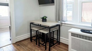 A small room with a wooden floor, a white wall, a TV mounted above a table with two chairs, and a window with curtains. - Hamilton Hideaway (Hagerstown, MD) (Hagerstown)