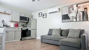 Une cuisine moderne avec des meubles blancs, un four à micro-ondes, une machine à café et une cuisinière. Le salon est doté d’un canapé gris agrémenté de coussins à motifs.