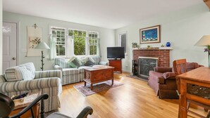 A living room with a sofa set, a wooden coffee table, a TV, a fireplace, and a dining table with chairs. - The Grounds - Brewster, MA (Brewster)