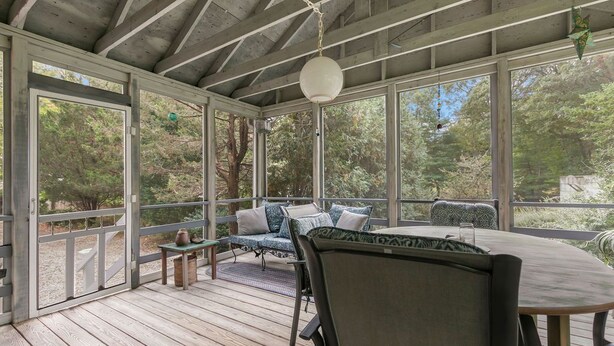 A covered outdoor seating area with a table and chairs, surrounded by large windows offering views of trees. - The Grounds - Brewster, MA (Brewster)