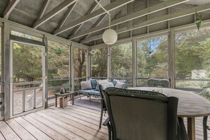 A covered outdoor seating area with a table and chairs, surrounded by large windows offering views of trees.