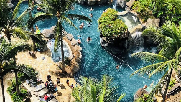 Un resort tropical con una gran piscina rodeada de palmeras y formaciones rocosas.