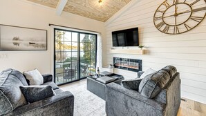 A modern living room with a large wall clock, a fireplace, and a large window overlooking a scenic view. - Canoes & Dock: Waterfront Retreat on Lake Beulah! (East Troy)