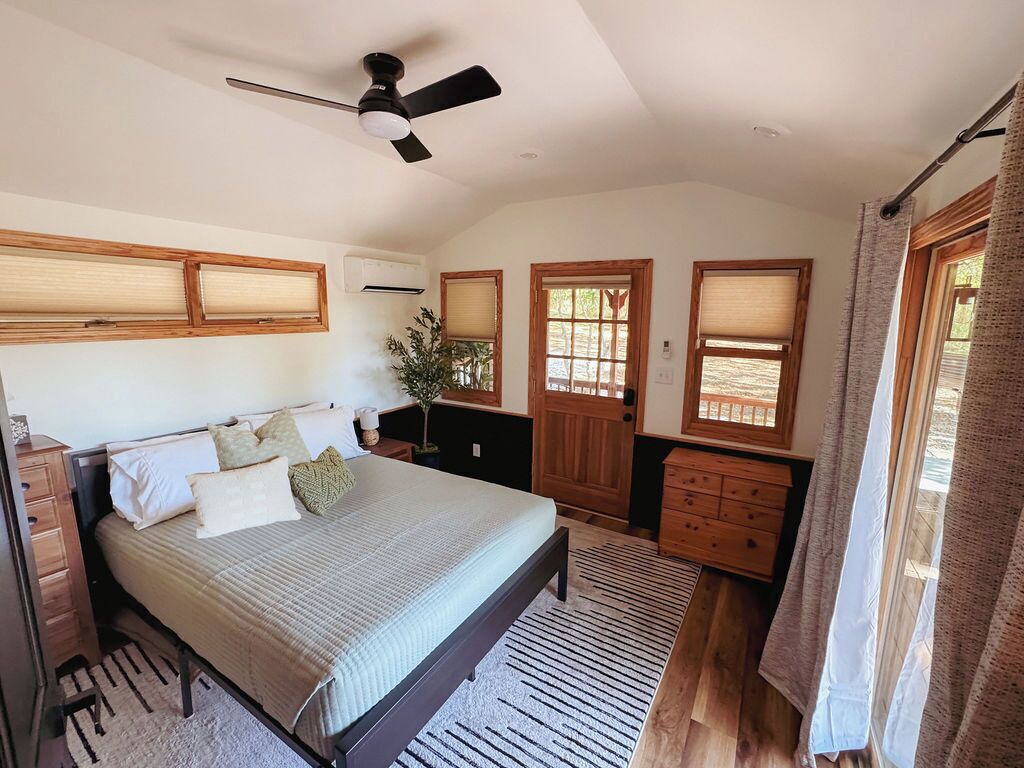 Ein Schlafzimmer mit Bett, Deckenventilator, Holztüren, Kommode und einem Fenster mit Vorhängen.