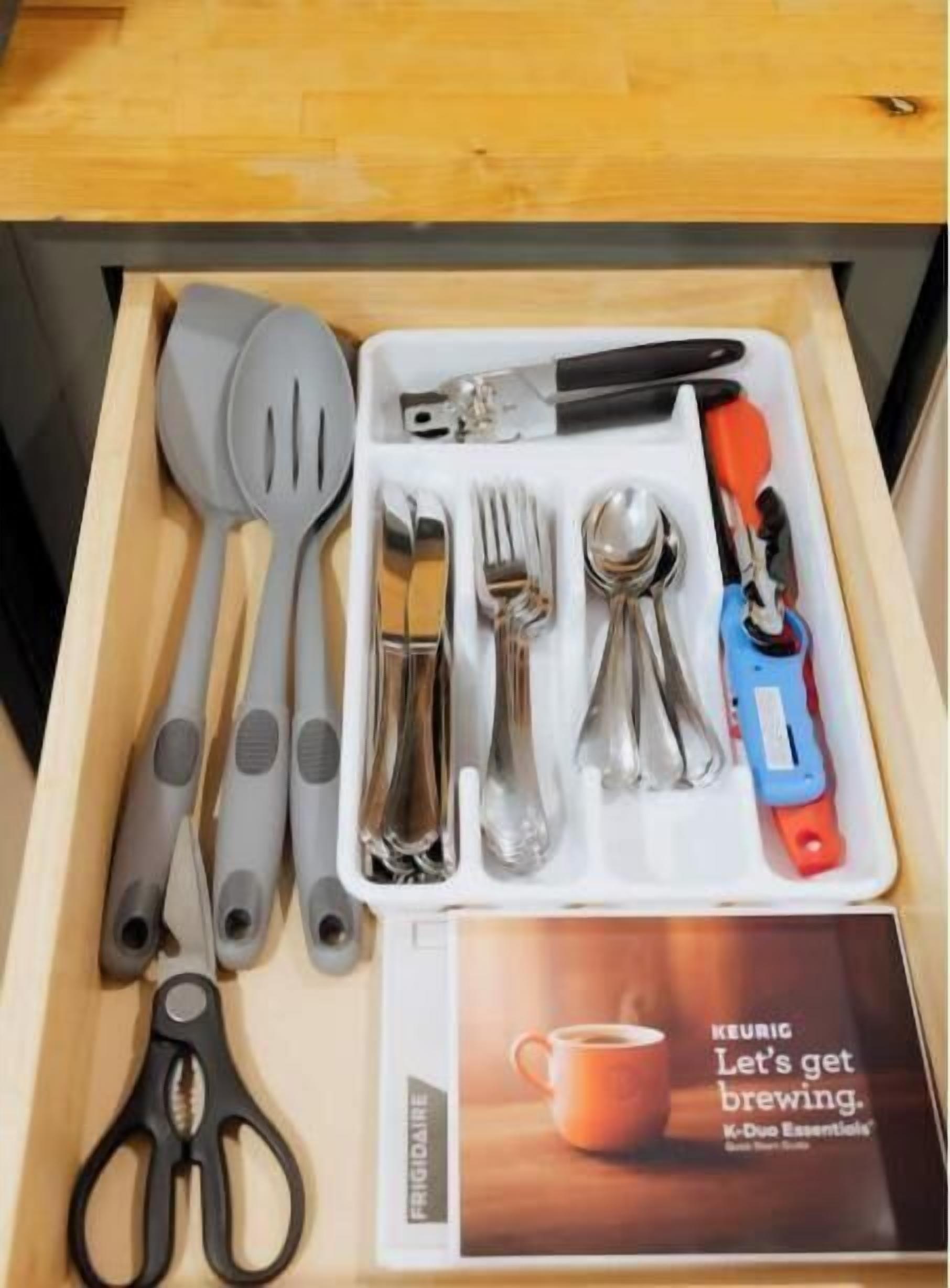Un tiroir contenant un ensemble d’ustensiles de cuisine, notamment une spatule, des pinces, un couteau, une cuillère, une fourchette, une paire de ciseaux et une machine à café.