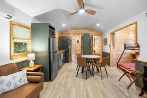 A compact kitchen with a refrigerator, microwave, and wooden cabinets. There is a dining area with a table and chairs, and a small seating area with a sofa and a lamp. - The Narrows Cabins #2964 (Marshall)