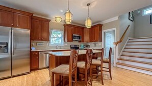 A kitchen with wooden cabinets, stainless steel appliances, a central island with chairs, pendant lighting, and a staircase to the right.