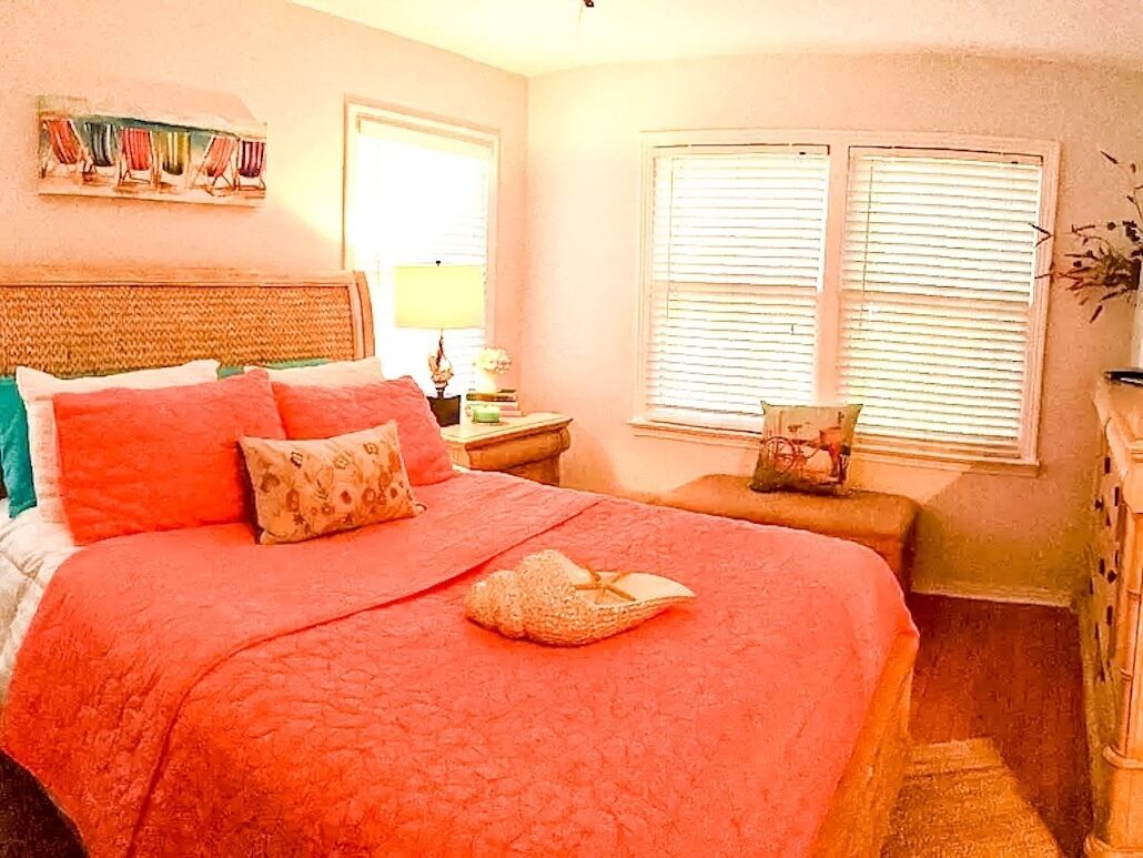 A bedroom with a large bed, a nightstand with a lamp, a window with blinds, and a decorative item on the nightstand.