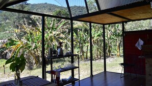 A room with large windows, a table with a vase of flowers, and a shelf with kitchen items. - Araucaria Cabin: Design and Nature (Itajubá)