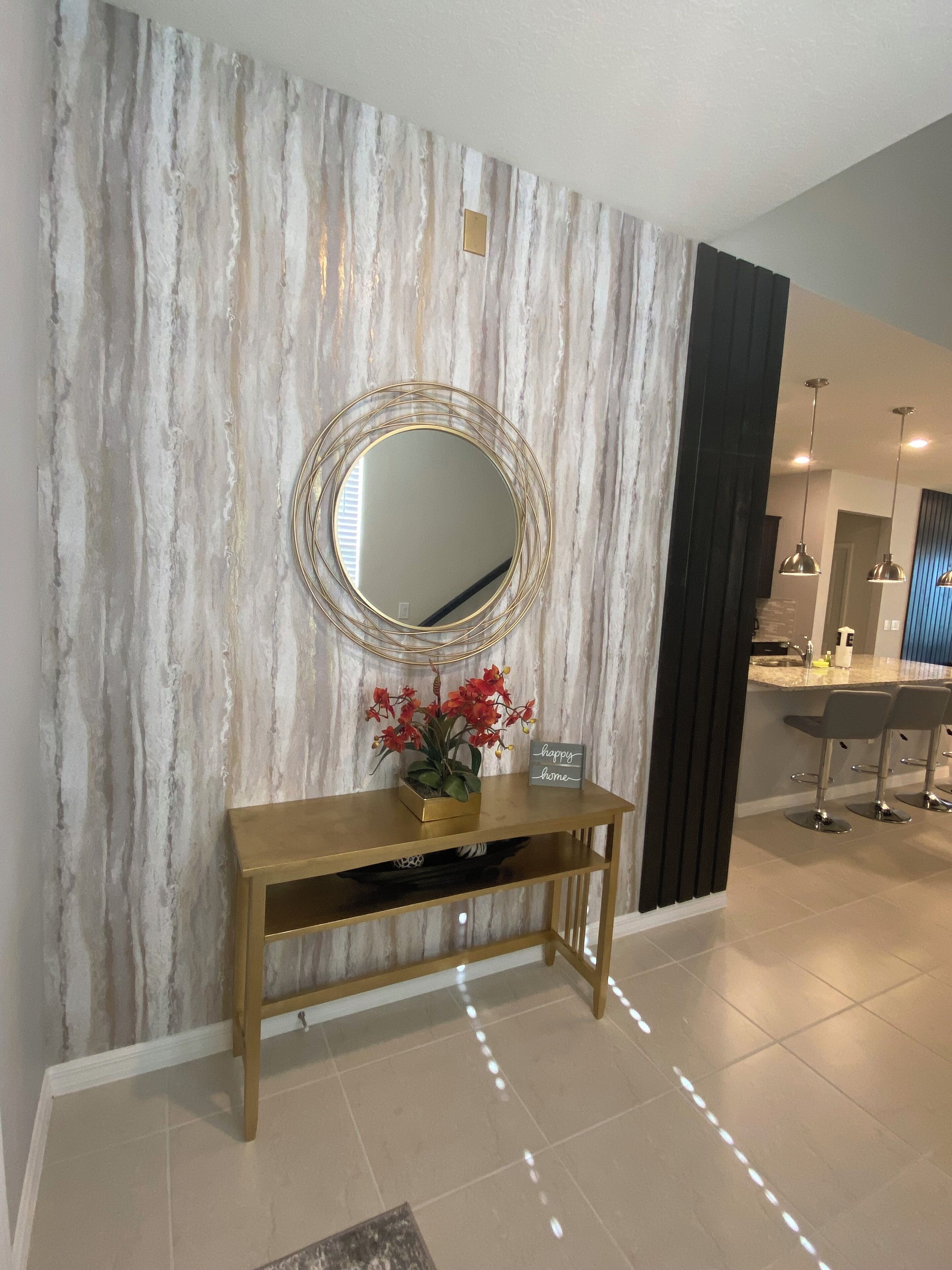 Un intérieur moderne avec un mur à motifs de marbre, un miroir rond, une console en bois sur laquelle est posée une plante en pot, et un coin cuisine à l’arrière-plan.