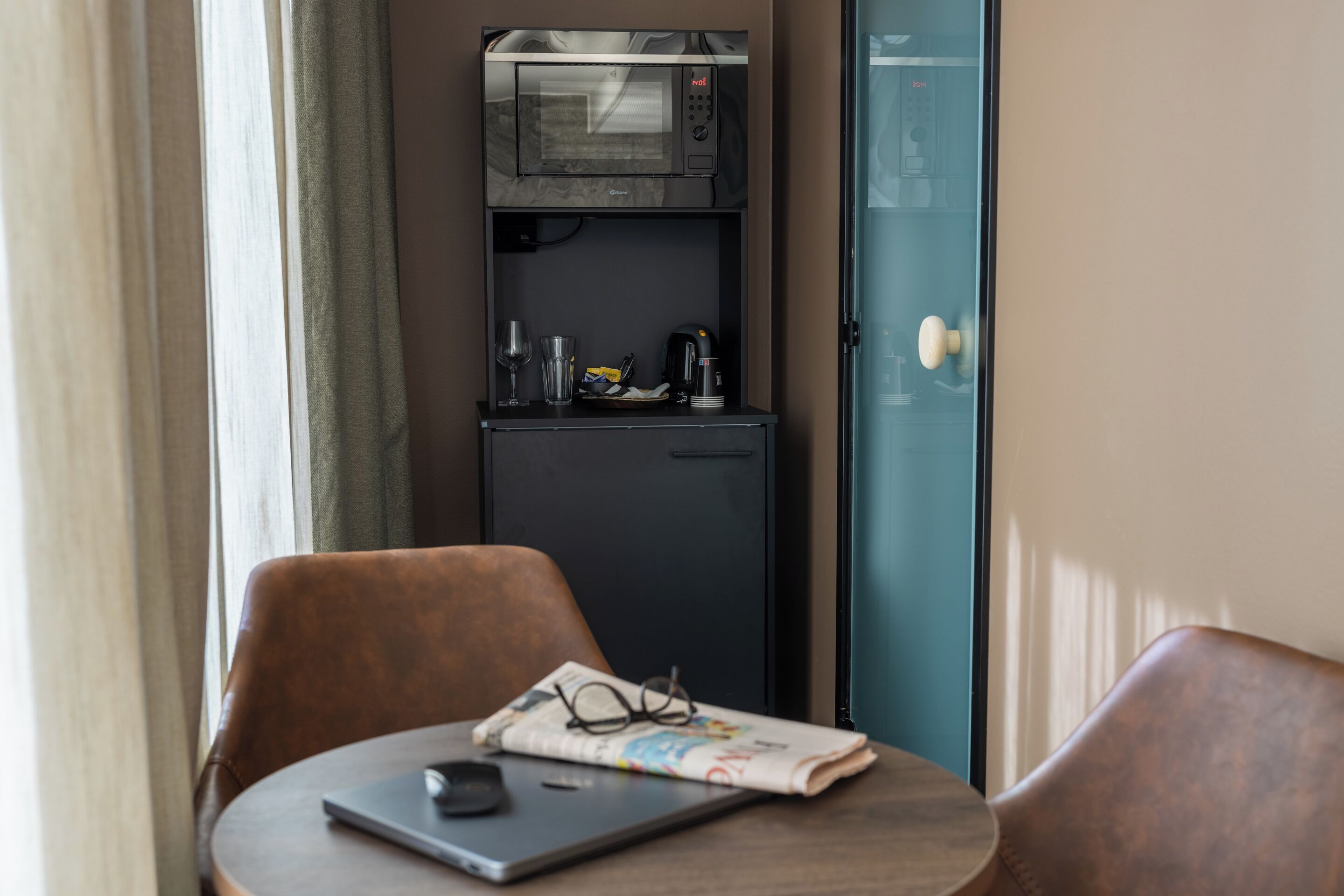 A compact kitchenette with a microwave, a small refrigerator, and a coffee maker. There is a laptop and a pair of glasses on a table.