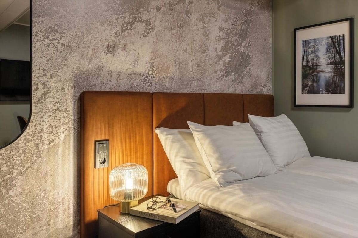 A bedroom with a wooden headboard, white bedding, a bedside table with a lamp, and a framed picture on the wall.